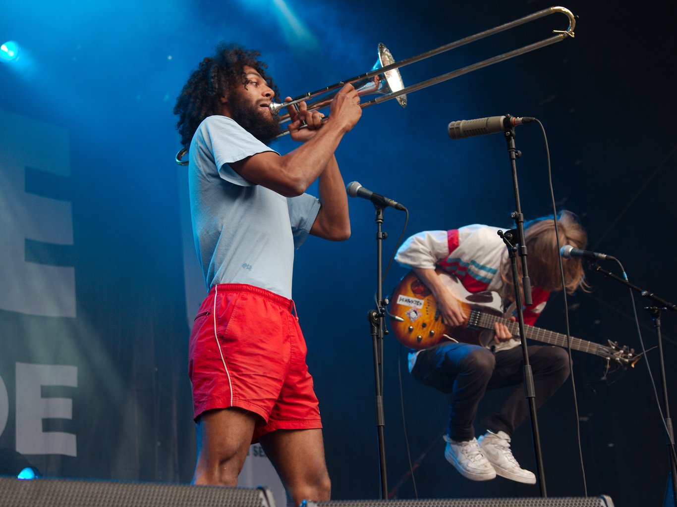 Guy in foreground playing a trombone, guitarist in background is in the middle of a jump, legs drawn to his body