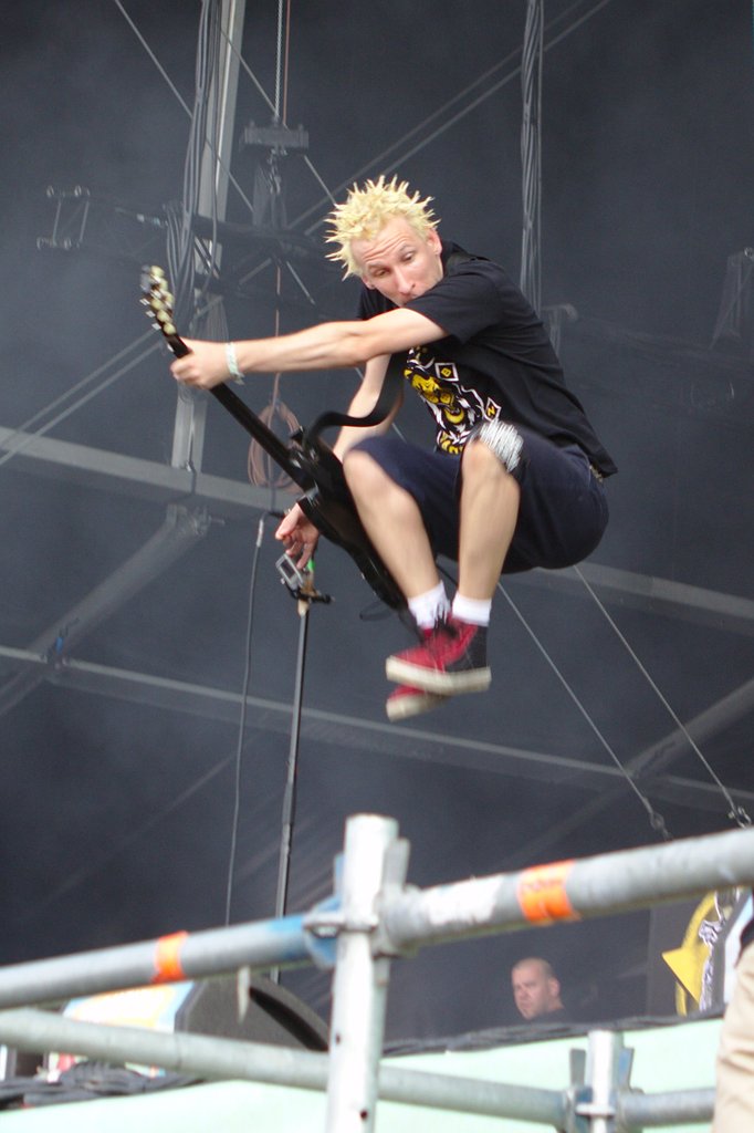 ZSK guitarist with yellow spiky hair is in the middle of a jump, legs drawn close to his body
